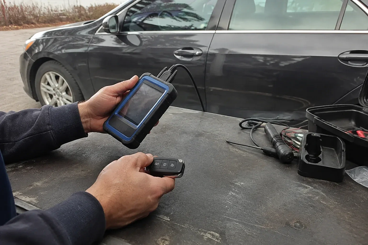 Hands holding a blue-edged electronic device and car-key fob at a worktable beside a parked car. — Transponder Key Programming, Markham