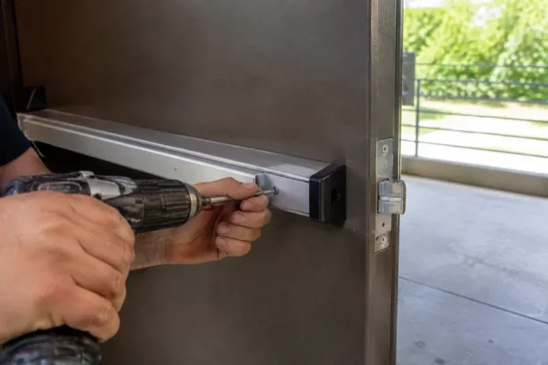 Hands using a power driver at the end of a horizontal push bar on an open metal door. — Panic & Exit Hardware, Markham